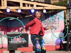 Artista de circo com maquiagem artística e figurino colorido segurando dois grandes aros de bambolê (hula hoops) azuis em um palco ao ar livre. Ao fundo, faixa com os dizeres "Transversidade" e iluminação de refletores.