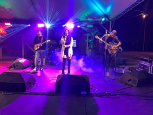 Banda ao vivo com vocalista feminina e dois guitarristas masculinos tocando em um palco ao ar livre com iluminação de palco vibrante rosa, azul e roxa. À direita, outros músicos e equipamentos são visíveis.