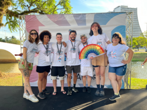 Grupo de sete pessoas em pé sorrindo em um palco ao ar livre, vestindo camisetas temáticas brancas, segurando um leque colorido com as cores do arco-íris e o texto "Mica Explica". Ao fundo, estrutura de palco e cenário com rio e ponte.