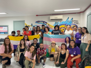 Amplo grupo diversificado de crianças, jovens e adultos reunidos assistindo a uma apresentação cultural ao ar livre, com artista fantasiado sentado no centro cercado pelo público sorridente.